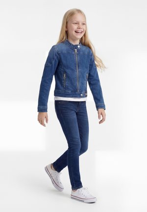 Mladé dievča kráča a usmieva sa, má na sebe modrú rifľovú bundu, modré džínsy, biele tenisky a biele tričko na jednoduchom pozadí.