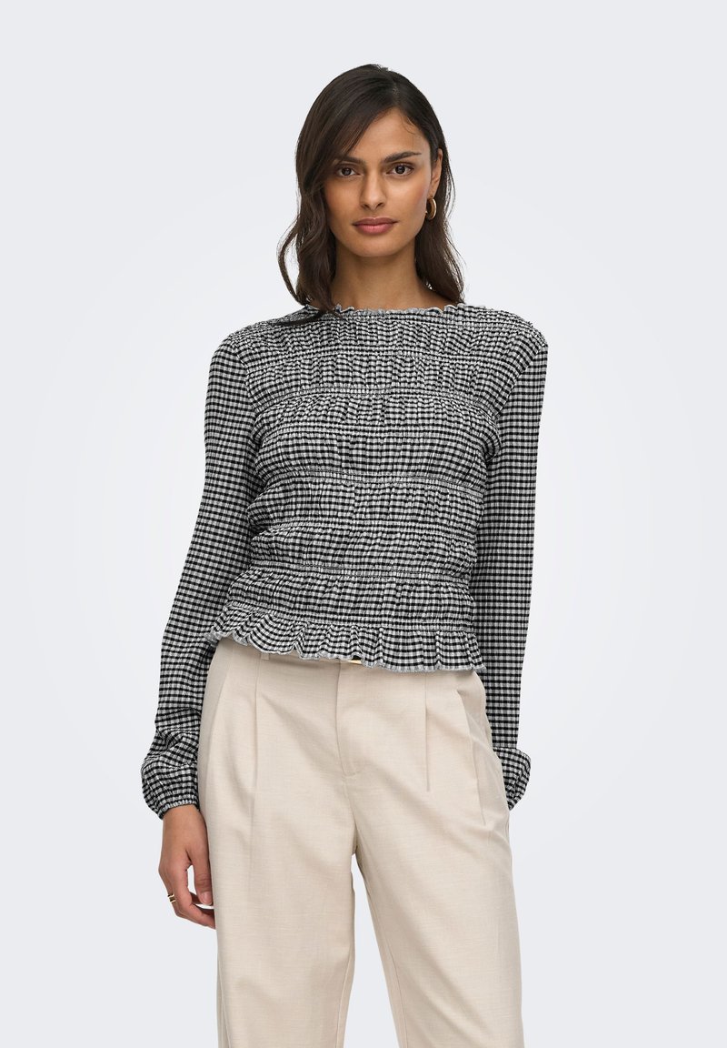 Femme portant un haut à manches longues à carreaux noir et blanc avec taille froncée et un pantalon beige taille haute, debout devant un fond uni.