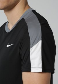 Sort atletisk T-shirt med grå og hvide skulderdetaljer. Har rund halsudsk æring, korte ærmer, glat stof og Nike-logo på fronten.