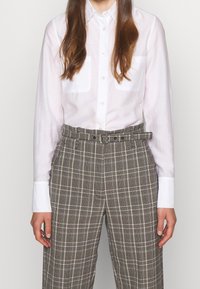 Vit långärmad skjorta med två bröstfickor; tvärs med hög midja, rutiga byxor i brunt och krämfärgat. Bältad design.