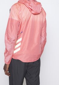 Leichter rosa Windbreaker mit strukturiertem Finish, ausgestattet mit einer Kapuze und diagonal verlaufenden weißen und farbigen Streifen auf dem Rücken.