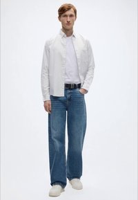 Λευκό πουκάμισο με λεπτές ρίγες και μακριά μανίκια, απλό λευκό μπλουζάκι από μέσα, φθαρμένο φαρδύ τζιν σε ανοιχτό γαλάζιο, μαύρη ζώνη, μπεζ αθλητικά παπούτσια.