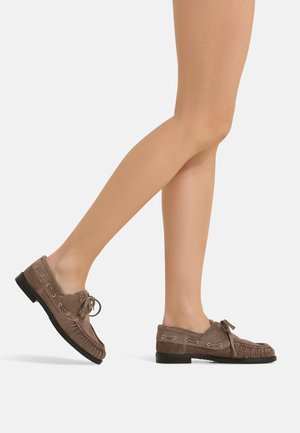 Legs wearing brown suede moccasin shoes with laces, one foot flat on the ground, the other lifted mid-step on a neutral background.