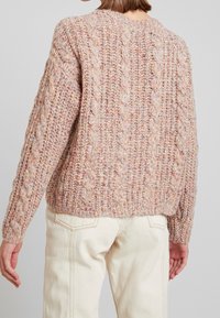 Femme portant un pull en maille épaisse aux tons pastel roses et un pantalon taille haute blanc cassé, vue de dos.