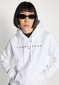 Felpa bianca con cappuccio caratterizzata dal logo "TOMMY JEANS" in nero, dotata di tasca frontale e stringhe. Abbinata a occhiali da sole neri e eleganti.