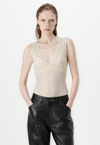 Pleteni brezrokavnik v svetli bež barvi z V-izrezom, ki ima vertikalne črte. Ujemajoče se z visokorasetimi črnimi usnjenimi hlačami s žepi.