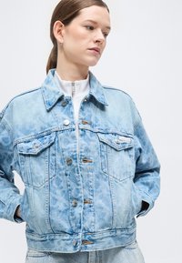 Γυναίκα που φοράει τζιν μπουφάν ανοιχτού μπλε με σχέδιο λεοπάρ, κλείνει με κουμπιά και έχει μπροστινές τσέπες, με τα χέρια στις τσέπες, κοιτάζει προς τα κάτω.