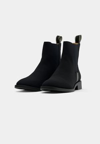 Paire de bottines noires à chevilles avec languettes, talons blocs bas et finition mate lisse sur fond blanc.