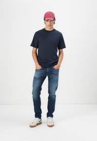 Marineblå t-skjorte, blå slitte jeans, hvite sneakers med brune såler, og en rød caps med svarte detaljer. Uformell antrekk på en modell.