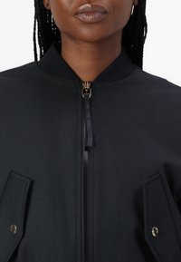 Zwarte bomberjack met een ritssluiting, puntige kraag, twee zijzakken en gouden hardwareaccenten, gemaakt van een gestructureerde stof.