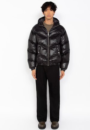 Chaquetón negro acolchado con una textura brillante, cierre de cremallera y capucha. Combinado con pantalones negros y calzado oscuro.