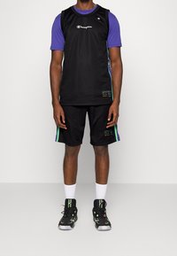 Muž oblečený v černém basketbalovém dresu Champion a šortkách, s fialovým tričkem pod ním, bílými ponožkami a černými vysokými sneakers s neonovými akcenty.