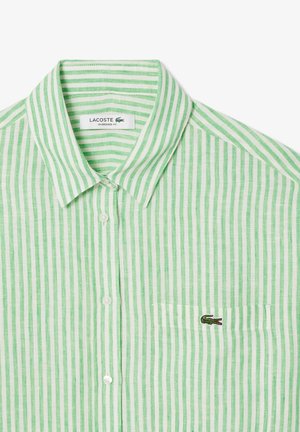 Camicia a righe verde e bianca con colletto, dotata di un piccolo logo del coccodrillo ricamato sulla tasca sinistra del petto. Taglio oversize.
