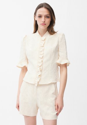 Junge Frau trägt cremefarbenes Kurzarmbluse mit Rüsche vorne und passende hoch taillierte Shorts vor weißem Hintergrund.