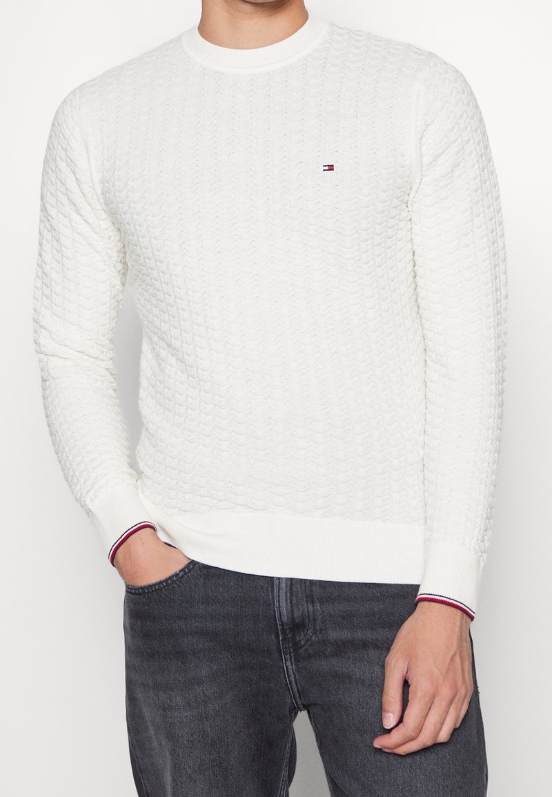 Pull en tricot blanc avec un motif texturé, col rond et poignets côtelés, arborant des rayures rouges et marines. Petit logo sur la poitrine.