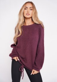 Sweter w kolorze śliwkowym, dziany, z szerokimi rękawami, regulowanym ściągaczem u dołu i miękką teksturą, noszony z czarnymi legginsami.