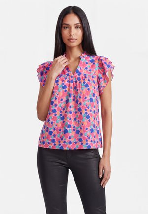 Vrouw met lang donker haar die een kleurrijk roze bloemige korte mouwen blouse en zwarte broek draagt, staand tegen een witte achtergrond.
