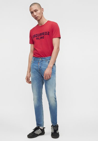 Άνδρας που φοράει κόκκινο t-shirt "DSQUARED2 MILANO", ανοιχτό μπλε τζιν και μαύρα αθλητικά παπούτσια με λευκές ρίγες, στέκεται μπροστά σε απλό ανοιχτό φόντο.