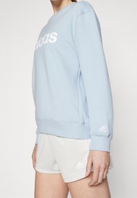 Light blue sweatshirt with crew neck, white "adidas" logo on front, cuffed sleeves, shown with light gray shorts featuring logo.