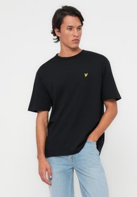 Camiseta negra de manga corta hecha de algodón, con un pequeño emblema de águila amarilla en el lado izquierdo del pecho, combinada con jeans de color azul claro.