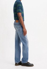 Man med en blå och grön randig kortärmad polo, ljusblå raka jeans och svarta läderskor, står på sidan mot vit bakgrund.