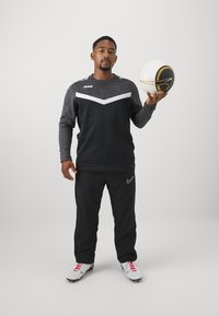 Atleta in una maglietta a maniche lunghe nera e grigia con un motivo a zigzag bianco, pantaloni neri e scarpini bianchi con accenti rosa, che tiene in mano un pallone da calcio a fantasia.