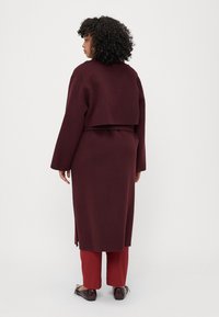 Burgundy coat with a cropped design, long sleeves, and side slits, paired with red pants and glossy shoes, viewed from the back.