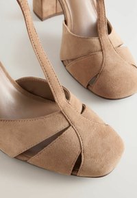 Zapatos de tacón alto de ante beige con tiras cruzadas, puntera redondeada y diseño recortado. El tacón grueso ofrece estabilidad y estilo.