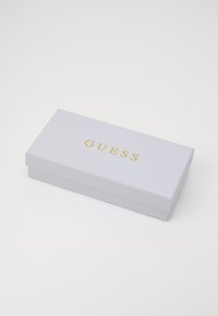 Scatola regalo rettangolare bianca con superficie texturizzata, con "GUESS" impresso in oro sulla parte superiore. Si chiude con un coperchio staccabile.