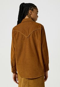 Femme avec des cheveux tressés portant une chemise en velours côtelé marron et une jupe assortie, de dos contre un fond uni clair.