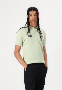 Šviesiai žalia polo marškinėlių su apykakle, su juodu "ellesse" logotipu ir spalvingu emblema. Juodos cargo kelnės.