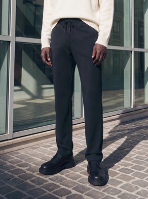 Personne portant un pantalon noir à cordon, des chaussures noires à lacets et un pull en maille blanc debout sur des pavés près de fenêtres en verre.