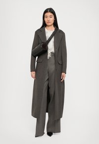 Woman wearing a long gray coat, wide-leg pants, a white textured top, black pointed shoes, and a black cylindrical crossbody bag.