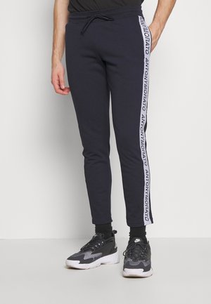 Man draagt zwarte joggerbroek met witte zijstrepen van het merk "ANTONYMORATO" en zwarte en grijze Nike sneakers.