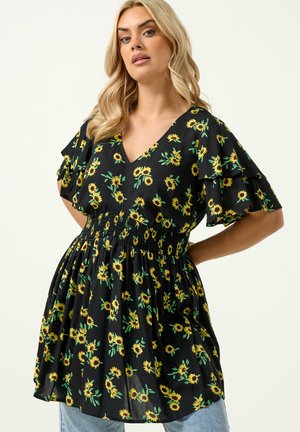 Lyshåret kvinde iført sort kjole med lagdelte ærmer og solsikkeprint, poserer med hænder på hofterne mod en ensfarvet baggrund.