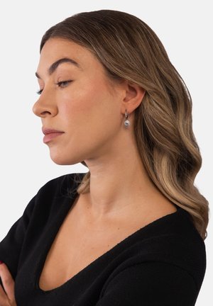 Woman with light brown wavy hair wearing a black V-neck top and silver hoop earrings with a detailed charm, facing left in a neutral background.