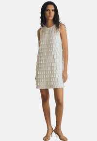 Valge ilma varrukateta kleit, millel on kihiline disain ristkülikukujuliste kangast klappidega, tekstureeritud ülaosa ja sirge lõige reie keskmise pikkuseni.