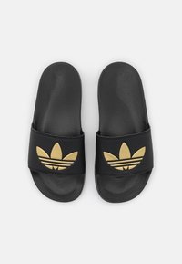 Svarta Adidas flip-flops med texturerade fotbäddar och gyllene Trefoil-logotyper på breda remmar, visade uppifrån mot en vit bakgrund.