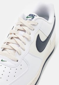 Baskets blanches Nike Air Force 1 avec lacets beiges texturés, logo swoosh noir, détails verts brodés et boîte à orteils perforée.
