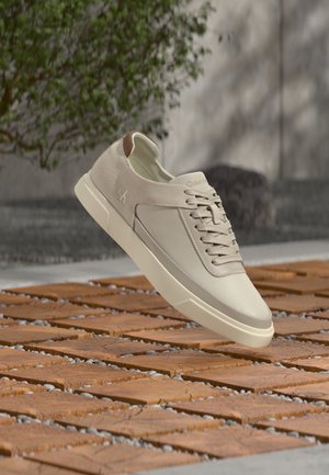 Baskets beige clair avec lacets flottant au-dessus d’un sol en carreaux de bois, cadre extérieur avec feuillage vert et mur en béton gris en arrière-plan.