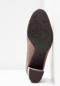 Semelle de chaussure en daim marron avec motifs de pièces embossées, nom de la marque "KAISER", et talon bloc reposant contre un mur blanc.