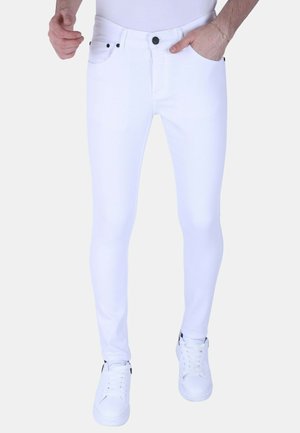 Man draagt strakke witte jeans, witte sneakers en een wit overhemd, met één hand in de zak en de andere ontspannen langs het lichaam.