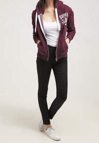 Sudadera con capucha de cremallera en color granate con forro gris, detalle de logo; camiseta sin mangas blanca; pantalones ajustados negros; zapatillas blancas. Conjunto informal en general.