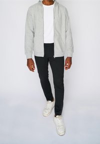 Felpa grigia con zip sopra una T-shirt bianca, abbinata a pantaloni neri slim-fit e sneakers bianche con una texture consumata.