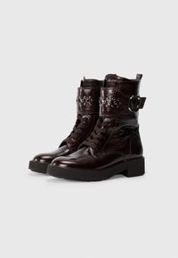 Brown patent leather ankle boots with a chunky sole, lace-up front, decorative studs, and an adjustable strap feature.