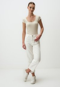 Body beige a costina con scollatura a forma di cuore, abbinato a pantaloni bianchi a vita alta e sneakers bianche, esposto su uno sfondo neutro.