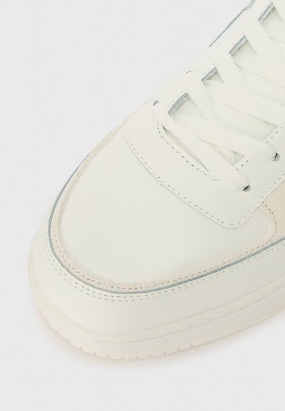 Λευκό δερμάτινο αθλητικό παπούτσι με στρογγυλή μύτη, υφή σ�ουέτ σε διάφορες περιοχές, επίπεδα κορδόνια και ορατές ραφές κατά μήκος των άκρων.