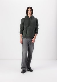 Sweatshirt cu glugă de culoare verde măsliniu, cu un buzunar frontal, asortat cu blugi gri decolorat și pantofi negri, pe un fundal alb simplu.