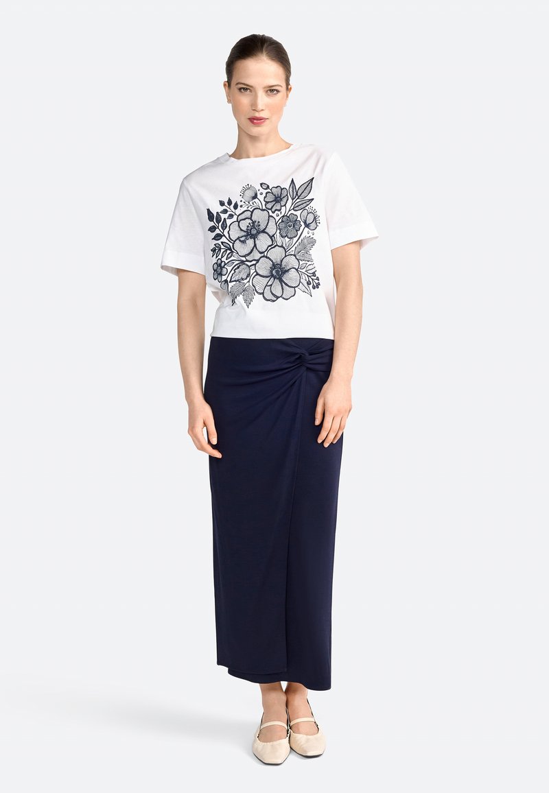 Witte katoenen t-shirt met marineblauwe bloemenprint, gecombineerd met een marineblauwe, knielange wikkelrok met een gedraaid detail aan de voorkant. Beige platte schoenen.