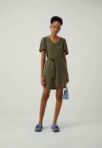 Vestido curto verde-caqui com mangas bufantes curtas, decote em V e um cinto a condizer. Combinado com sandálias azuis e uma pequena bolsa azul.